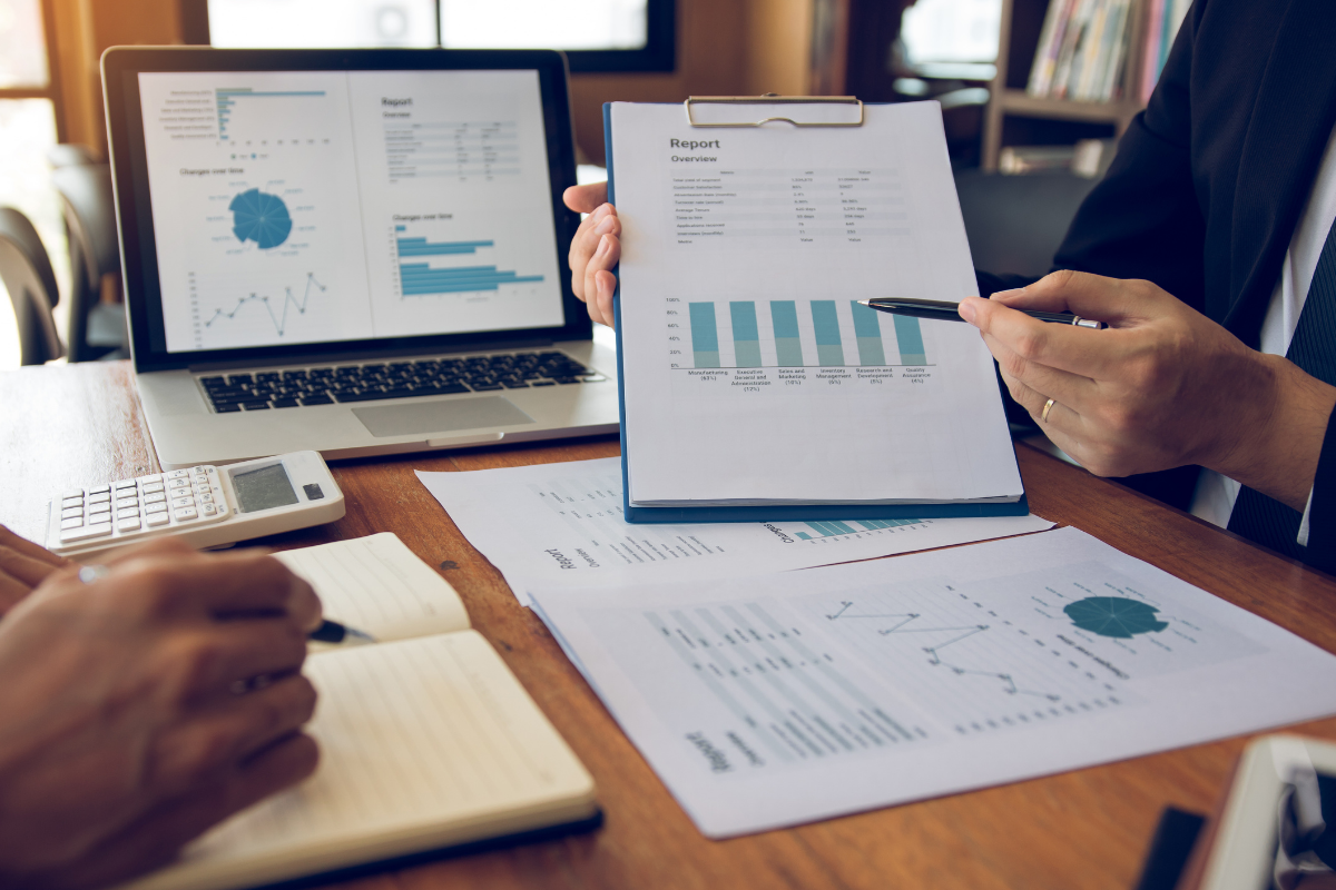 Accoutant's hand pointing towards charts on a report attached to a clipboard while at a desk.