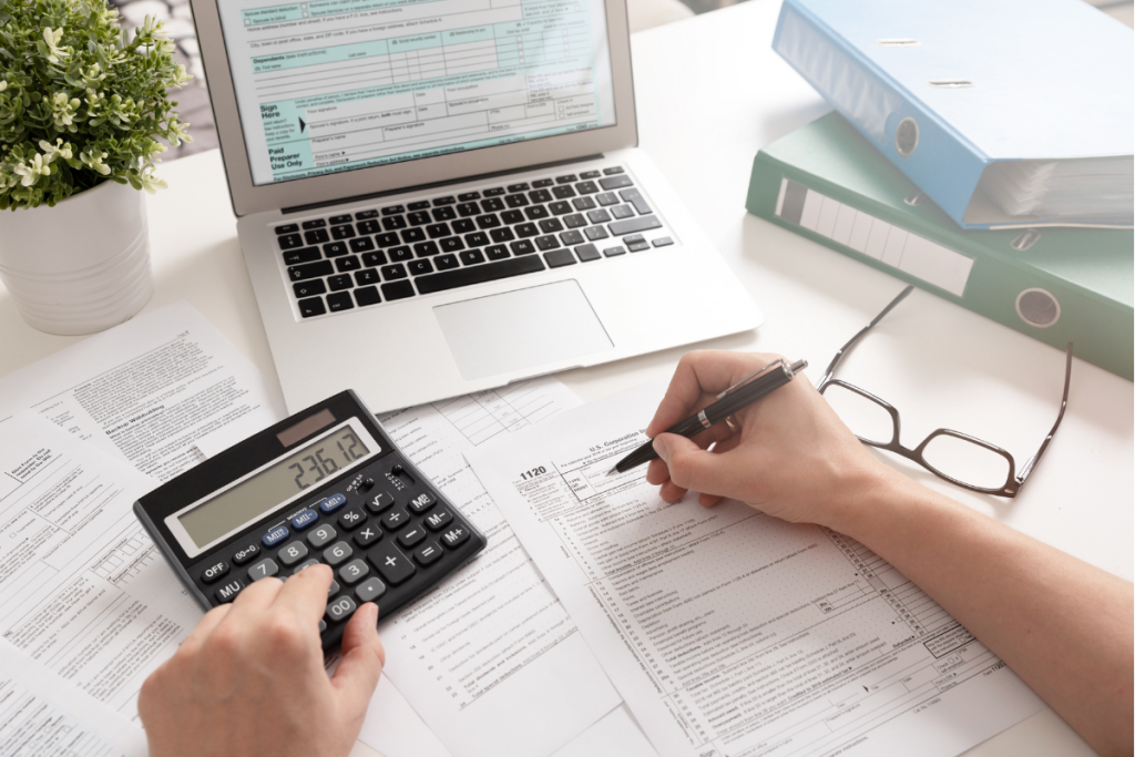 Point of view, accountant holds pen while typing figures into a calculator, laptop in the background - Tax Accountants Essex