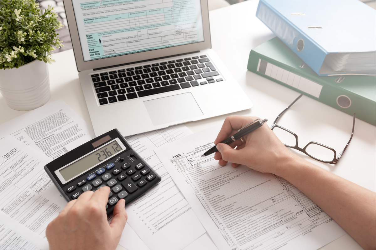 Point of view, accountant holds pen while typing figures into a calculator, laptop in the background